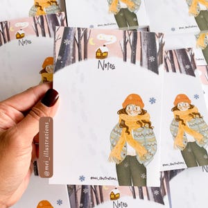 Puede incluir: Un montón de tarjetas de notas con temática invernal. Cada tarjeta presenta una ilustración de una persona en una escena invernal, con gorro y bufanda naranja. La palabra "Notes" está escrita en cada tarjeta. Las tarjetas son blancas con un fondo de bosque invernal.
