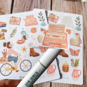 May include: A close-up of a white marker holding a sticker of a vintage typewriter. The sticker is peach-colored with a detailed design. Other stickers with similar illustrations are in the background, including a person riding a bicycle and floral elements.
