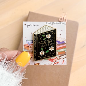 Könnte beinhalten: Ein schwarzer Emaille-Pin in Form eines offenen Buches mit goldenem Rand. Der Pin zeigt den Text "this too shall pass" und Gänseblümchen- und Blattdesigns. Der Pin befindet sich auf einer weißen Karte mit dem Text "gentle reminder".