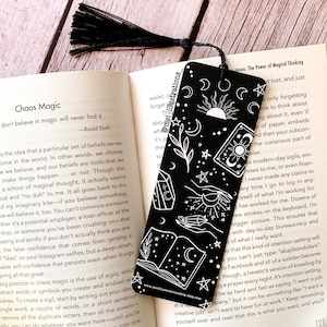 Może przedstawiać: Czarna zakładka do książki z białymi niebiańskimi i mistycznymi ilustracjami, w tym księżycami, gwiazdami, słońcem, kryształami i kartami tarota. Zakładka ma czarny frędzel i jest umieszczona na otwartej książce. Tekst na zakładce brzmi "@meiillustrationshop.etsy.com".