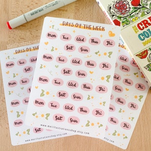 Può includere: Tre fogli di adesivi con la scritta "DAYS OF THE WEEK" e i giorni della settimana in un ovale rosa. Gli adesivi sono decorati con piccoli fiori e cuori. Un pennarello e un libro da colorare sono sullo sfondo.