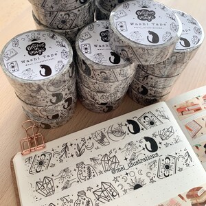 May include: Several rolls of black and white washi tape with a celestial design, including moons, stars, crystals, and a black cat. The tape is labelled "Believe in Magic" and "Washi Tape". A notebook with washi tape samples and a rose gold clip are also visible.