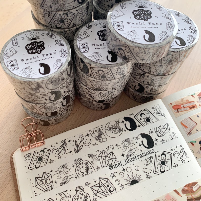 May include: Several rolls of black and white washi tape with a celestial design, including moons, stars, crystals, and a black cat. The tape is labelled "Believe in Magic" and "Washi Tape". A notebook with washi tape samples and a rose gold clip are also visible.