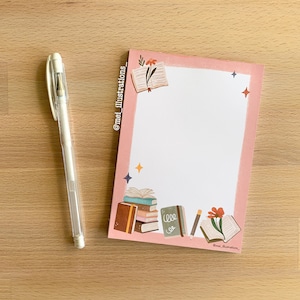 May include: A white gel pen lies next to a pink notepad with a decorative border. The notepad features illustrations of books, flowers, and stars. The text "@mei_illustrations_" is visible on the notepad.
