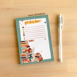 Puede incluir: Un bloc de notas turquesa y blanco con el texto "get stuff done" y una lista de líneas. Un bolígrafo blanco descansa junto al bloc de notas. El bloc de notas presenta ilustraciones de libros y una persona leyendo. El bloc de notas está sobre una superficie de madera.