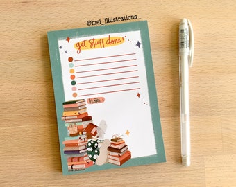 Könnte beinhalten: Ein türkis-weißes Notizblock mit dem Text "get stuff done" und einer Liste von Zeilen. Ein weißer Stift liegt neben dem Notizblock. Der Notizblock zeigt Illustrationen von Büchern und einer lesenden Person. Der Notizblock liegt auf einer Holzoberfläche.