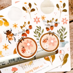 May include: A collection of spring-themed stickers featuring floral and insect designs. The stickers include illustrations of ladybugs, flowers, and strawberries. The text "120gr smooth matte" and "Usual eco white matte paper" are visible. A marker and the text "Spring Vibes Vol. 2" are also present.