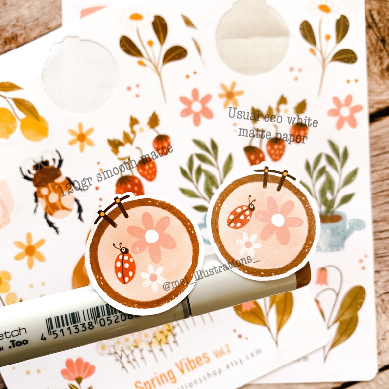 May include: A collection of spring-themed stickers featuring floral and insect designs. The stickers include illustrations of ladybugs, flowers, and strawberries. The text "120gr smooth matte" and "Usual eco white matte paper" are visible. A marker and the text "Spring Vibes Vol. 2" are also present.