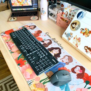 May include: A workspace setup featuring a laptop, keyboard, and mouse on a floral-patterned desk mat. The mat and laptop have illustrations of women. A water bottle and other desk accessories are also visible.