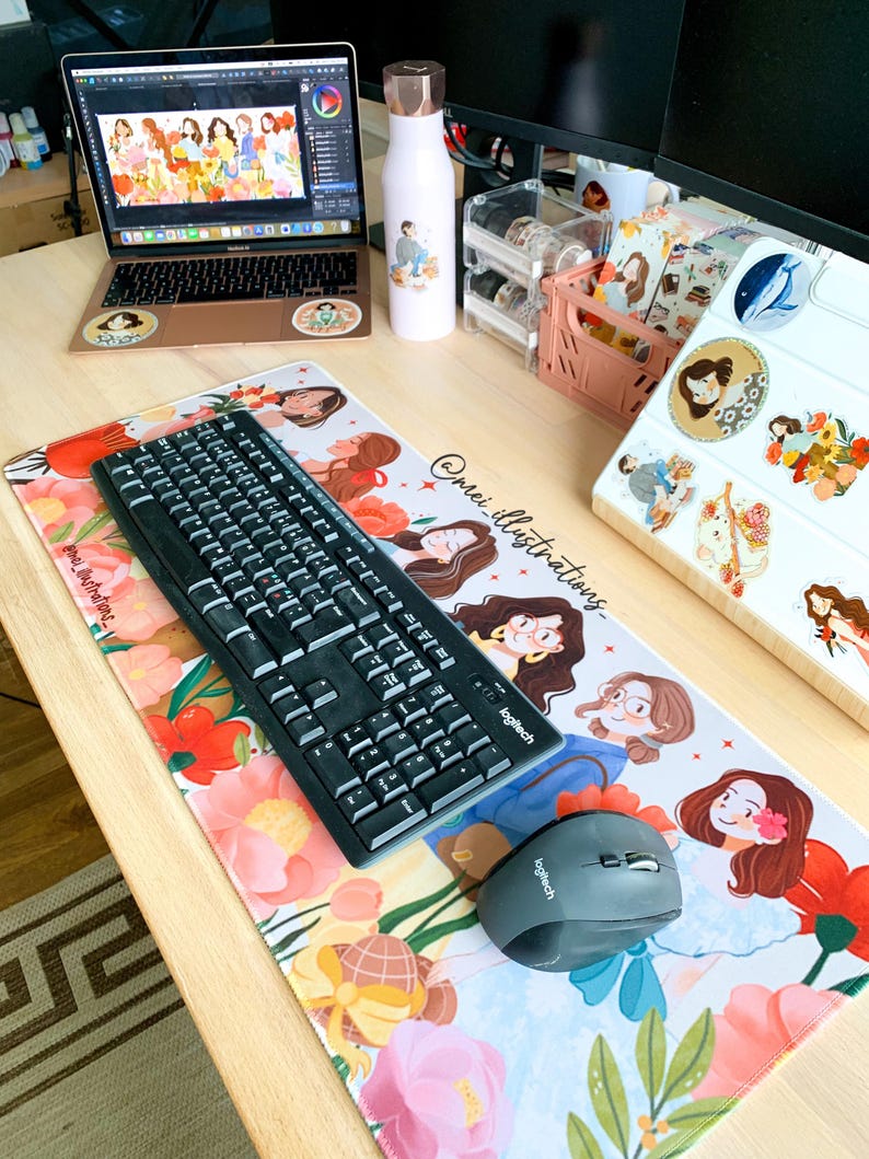 May include: A workspace setup featuring a laptop, keyboard, and mouse on a floral-patterned desk mat. The mat and laptop have illustrations of women. A water bottle and other desk accessories are also visible.