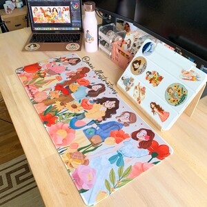 May include: A desk setup featuring a floral-themed mousepad with illustrations of women, a laptop, a water bottle, and a tablet with stickers. The mousepad has the text "@mei_illustrations". The color palette includes pink, orange, and yellow.