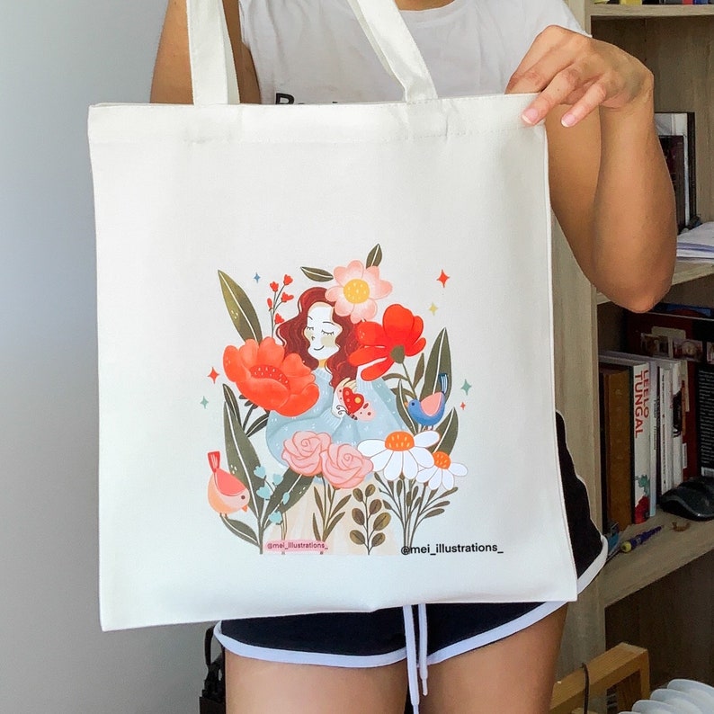 May include: A cream-coloured tote bag with a vibrant illustration of a woman amidst flowers and birds. The design includes red poppies, pink roses, and daisies. The artist's name, "@mei_illustrations_," is printed at the bottom of the bag.