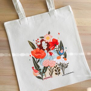 May include: Off-white tote bag with a vibrant illustration of a woman amidst flowers and birds. The design includes red poppies, pink roses, and a blue bird. The artist's name, "mei_illustrations_," is printed on the bag.