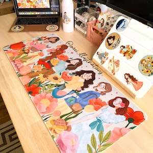 May include: A colorful desk setup featuring a laptop, water bottle, and a large mousepad. The mousepad displays an illustration of several women surrounded by flowers. The water bottle and laptop have matching illustrations. The text "@mei_illustrations" is visible.