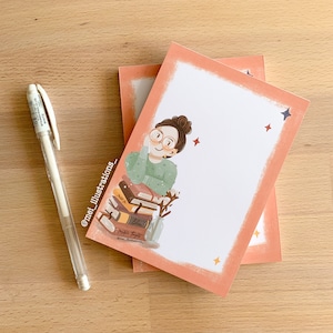 Op de afbeelding: Twee notitieblokken met een perzikkleurige rand en een cartoonillustratie van een persoon met een bril, rustend op een stapel boeken. Een witte pen ligt op een houten oppervlak. De illustratie bevat de tekst "mei_illustrations_" en "Pretty Eight".