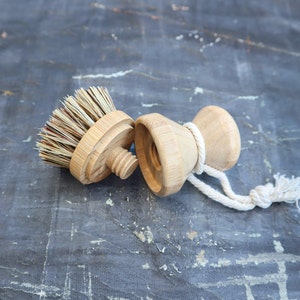 May include: A wooden brush with natural bristles and a wooden handle with a rope attached. The brush is designed for cleaning dishes or other surfaces.