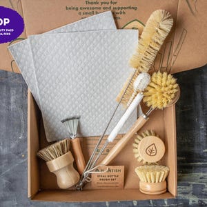 May include: A cleaning kit in a cardboard box containing two light gray textured cloths, a long-handled sisal bottle brush, a thin wire brush, a round wooden dish brush with a leaf icon, a small wooden scrub brush, and a small metal scraper.