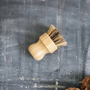 May include: A wooden brush with natural bristles, ideal for cleaning dishes or scrubbing surfaces. The brush has a light brown wooden handle and is lying on a grey textured surface.