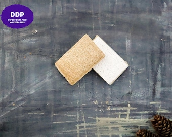 May include: Two rectangular sponges are positioned diagonally. One sponge features a tan, fibrous, textured surface, while the other sponge has a smooth, white, porous surface. The sponges are placed against a dark, mottled gray background.