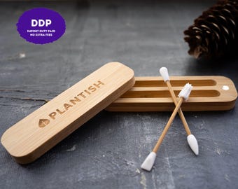 May include: A light brown bamboo storage case with the word PLANTISH and a leaf icon engraved on the lid. The case is open, revealing two internal slots. Two reusable cotton swabs with white silicone tips and bamboo stems rest on the case.