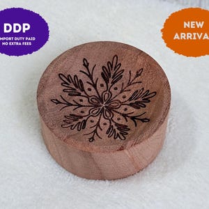 May include: A round, light brown wooden disc featuring a dark, laser-engraved snowflake design on its top surface. The wood grain is visible across the circular face, and the snowflake pattern consists of intricate, symmetrical branches.