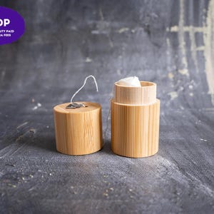 May include: A cylindrical bamboo dental floss container with a removable lid. The lid features a small metal cutter for the white floss, which is visible inside the open container. The natural light wood grain texture is clearly displayed on both parts.
