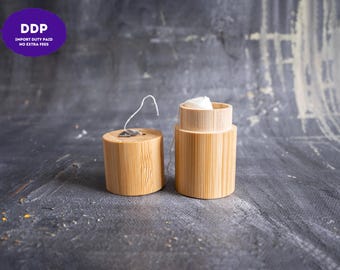 May include: A cylindrical bamboo dental floss container with a removable lid. The container is open, revealing a spool of white dental floss inside. A small metal cutting mechanism is attached to the top of the lid, with a piece of white floss extending from it.