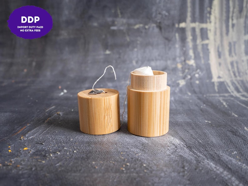 May include: A cylindrical bamboo dental floss container with a removable lid. The lid features a small metal cutter for the white floss, which is visible inside the open container. The natural light wood grain texture is clearly displayed on both parts.