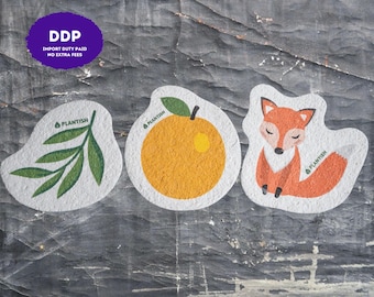 May include: Three white, irregularly shaped cleaning sponges featuring printed designs. One sponge displays a green leafy branch, the second shows an orange fruit with a green leaf, and the third depicts an orange fox. Each sponge includes the text PLANTISH.