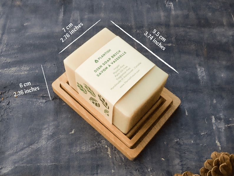 Pode incluir: Uma barra retangular de sab&atilde;o para lavar lou&ccedil;a em um prato de madeira. O sab&atilde;o &eacute; etiquetado como "PLANTISH DISH SOAP BRICK SAVON A VAISSELLE" e lista os ingredientes: "Vegano, &Aacute;gua, Azeite de oliva, &Oacute;leo de palma, Quinoa, Fragr&acirc;ncia, Mel". O sab&atilde;o mede 7 cm por 9,5 cm por 6 cm.