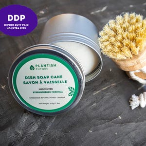 May include: A round silver metal tin containing a white dish soap cake with a green and white label reading Plantish Future Dish Soap Cake Savon à Vaisselle Unscented Strengthened Formula, accompanied by a round wooden dish brush with tan bristles.