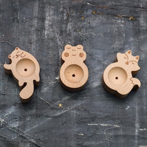 May include: Three wooden animal-shaped ornaments with a hole in the centre. The ornaments are a squirrel, a fox, and a bear. The ornaments are made of light brown wood.
