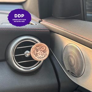 May include: A circular wooden car air freshener clip featuring a laser-engraved design of a hummingbird hovering near a flower, surrounded by small stars and leafy branches, attached to a circular silver-toned vehicle air vent.