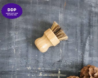 May include: A round wooden dish brush with stiff, light brown natural bristles. The handle is made of smooth, light-colored wood and is shaped to fit comfortably in the palm of a hand. The brush is designed for scrubbing dishes and surfaces.