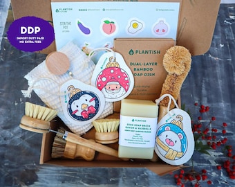 May include: A cardboard gift box containing a dual-layer bamboo soap dish, a dish soap brick, a long-handled sisal brush, a small round wooden brush, a textured white cloth, and three decorative holiday-themed tags featuring a penguin, Santa, and a bear.