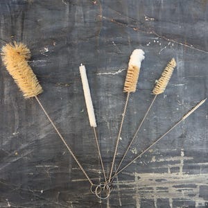 Peut inclure: Cinq brosses de nettoyage avec de longs manches fins et des poils naturels. Les brosses sont disposées en éventail sur une surface texturée grise.