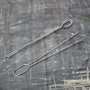 May include: Two silver metal tongs, one with a loop at the end and the other with a rounded end. The tongs are on a gray textured surface.