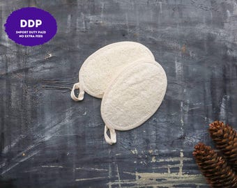 May include: Two oval-shaped, off-white loofah bath sponges with textured surfaces and fabric loops for hanging. The sponges have a natural, fibrous appearance and are positioned together, showcasing their rounded edges and woven trim.