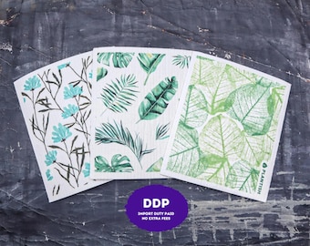 May include: Three rectangular cellulose sponge cloths featuring distinct botanical prints. The left cloth has light blue flowers on thin stems. The center cloth displays various green tropical palm leaves. The right cloth shows green leaf skeletons with the text PLANTISH.