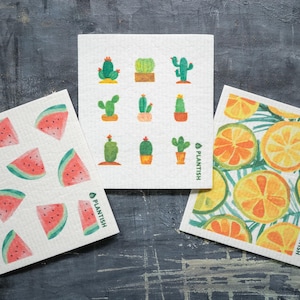May include: Three reusable cellulose sponges with different designs. The first sponge has a watermelon pattern with pink and green slices. The second sponge has a cactus pattern with green cacti in pots. The third sponge has a lemon pattern with yellow lemons and green leaves. All sponges have the word "Plantish" printed on them.