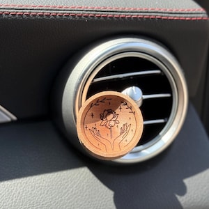 May include: A circular, wooden car air vent diffuser featuring a laser-etched floral design held by hands. The light brown diffuser is positioned in front of a silver air vent. The background shows a black car interior with red stitching.