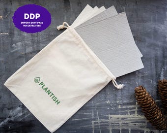 May include: A cream-colored cotton drawstring pouch with the green text PLANTISH and a leaf icon printed on the front. Inside the pouch are several square, light gray, textured sheets. Two brown pinecones are positioned near the pouch.
