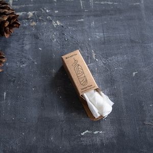 May include: A rectangular brown cardboard box containing white dental floss, with the text "PLANTISH" and "Dental Floss" printed on the front. The packaging features a simple line drawing of a floss container and is positioned near two pinecones.