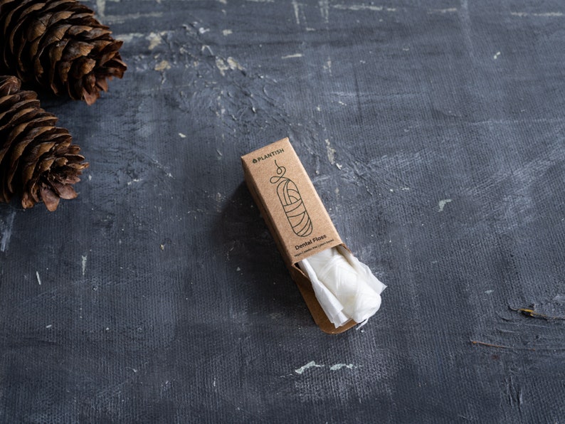 May include: A rectangular brown cardboard box containing white dental floss, with the text "PLANTISH" and "Dental Floss" printed on the front. The packaging features a simple line drawing of a floss container and is positioned near two pinecones.