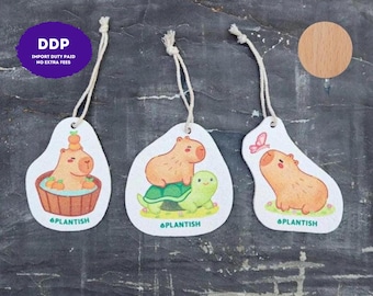 Puede incluir: Tres adornos colgantes blancos de forma irregular con ilustraciones de capibaras. Uno muestra un capibara en una bañera con naranjas, otro sobre una tortuga y otro con una mariposa. Cada uno tiene una cuerda y el texto PLANTISH.