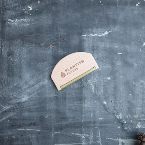 May include: A wooden fabric shaver with a gold metal blade. The shaver is half-circle shaped and has the words "Plantish Future" engraved on the front.