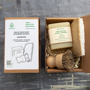 May include: A solid dish soap scrubber bundle in a cardboard box. The box contains a dish soap bar, a natural sponge, and a wooden handled brush. The box is labeled "Vegan, plastic-free, plant-based" and "Unscented".