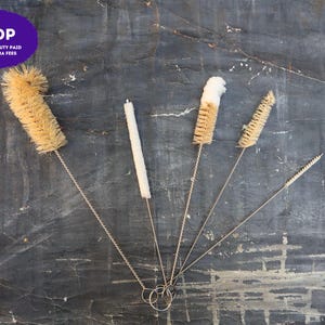 May include: A set of five cleaning brushes with twisted metal wire handles and varying bristle types, including natural tan fibers and white soft material. Each brush features a circular metal loop at the base for hanging and storage purposes.