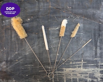 May include: A set of five cleaning brushes with twisted metal wire handles and varying bristle types, including natural tan fibers and white soft material. Each brush features a circular metal loop at the base for hanging and storage purposes.