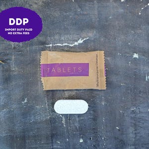 May include: A rectangular brown paper packet with a purple horizontal band labeled TABLETS and Multipurpose Cleaner, accompanied by a single white, oval-shaped speckled cleaning tablet positioned directly below the packet on a dark surface.